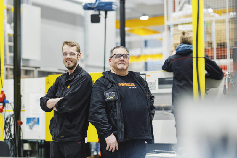 Kaksi mustaa univormua pukeutunutta miestä, mukaan lukien logolliset takit, seisovat selkä vastakkain hymyillen työpajassa. Koneet ja kirkkaan keltaiset aksentit näkyvät taustalla, mikä viittaa teolliseen ympäristöön.