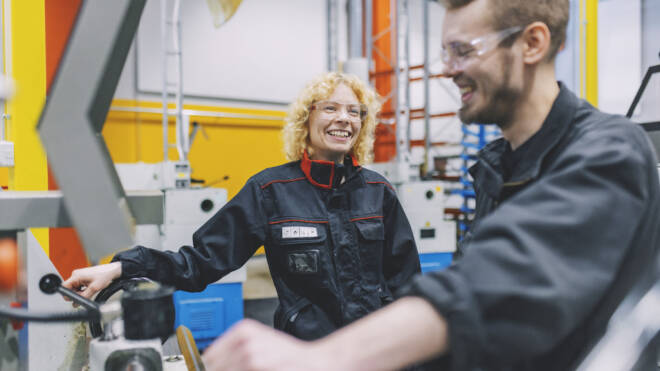 Kaksi henkilöä suojavarusteissa käyttää koneita teollisuusympäristössä. He hymyilevät ja näyttävät olevan mukana keskustelussa. Molemmat käyttävät suojalaseja ja haalareita, joiden taustalla näkyy erilaisia varusteita.