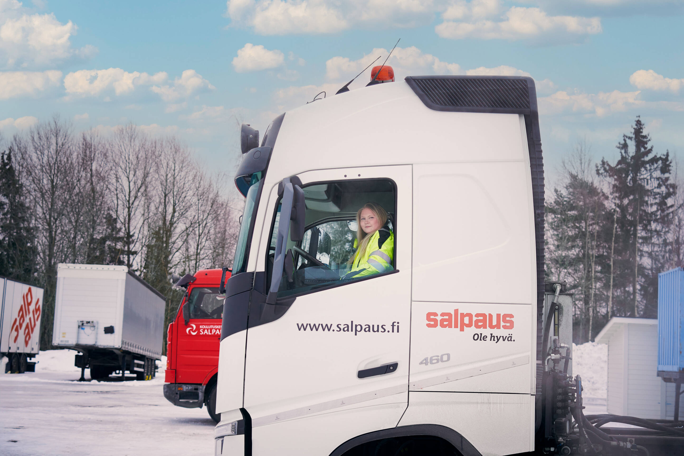 Keltaista turvaliiviä pukeutunut henkilö istuu valkoisen kuorma-auton kuljettajan paikalla, jonka sivulla on "salpaus.fi". Taustalla näkyy toinen punainen kuorma-auto, jossa on luminen maa ja lehdettömiä puita osittain pilvisen taivaan alla.