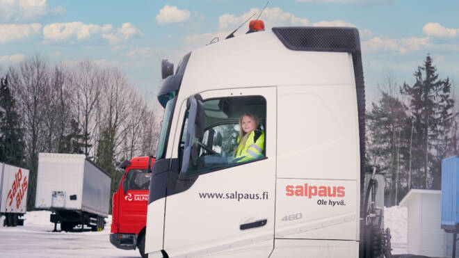 Keltaista turvaliiviä pukeutunut henkilö istuu valkoisen kuorma-auton kuljettajan paikalla, jonka sivulla on "salpaus.fi". Taustalla näkyy toinen punainen kuorma-auto, jossa on luminen maa ja lehdettömiä puita osittain pilvisen taivaan alla.