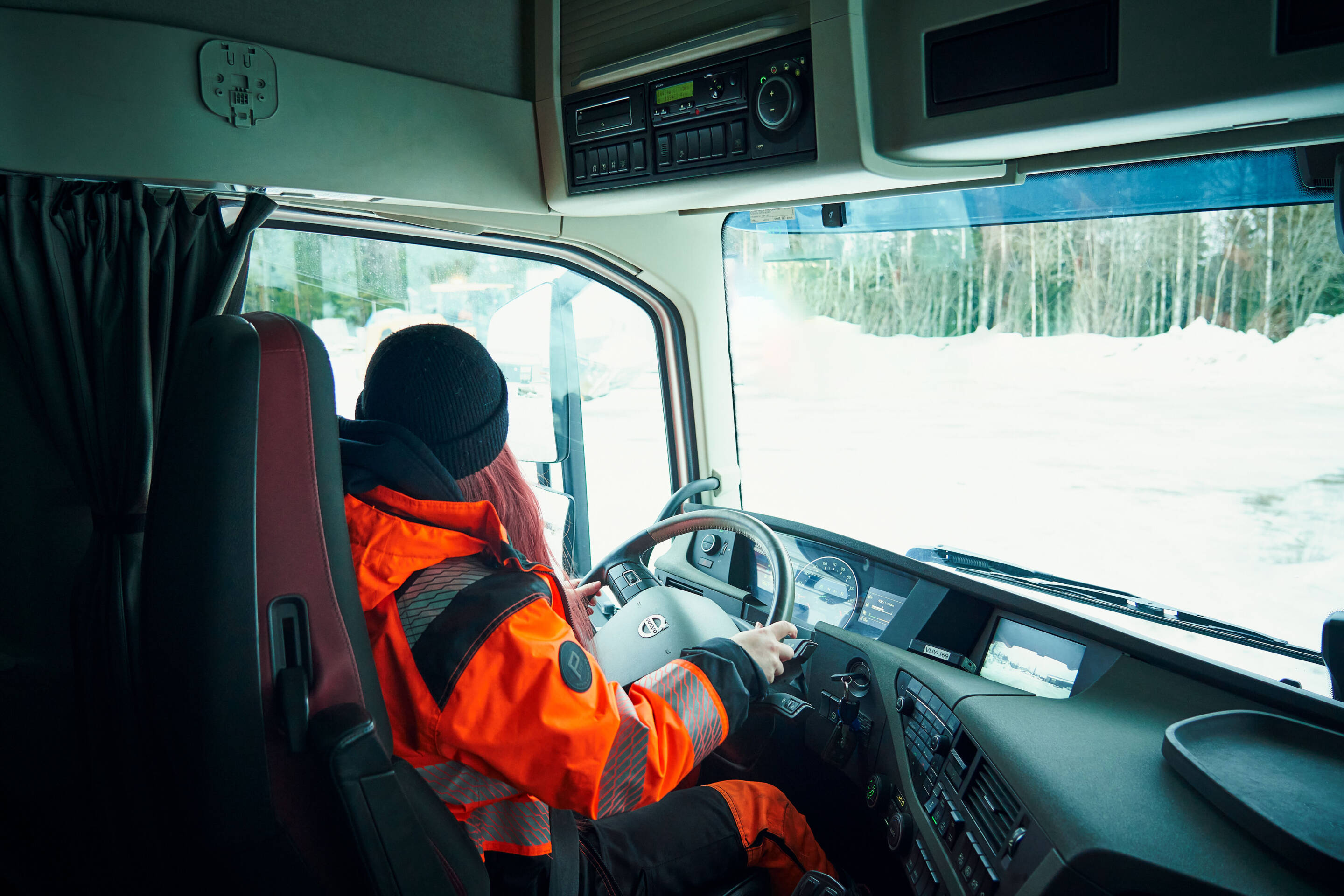 Kirkkaan oranssissa turvavarusteissa oleva henkilö ajaa suurta kuorma-autoa lumisessa maisemassa. Kojelaudassa on useita säätimiä, ja ulkonäkymässä näkyy puiden reunustama tie.