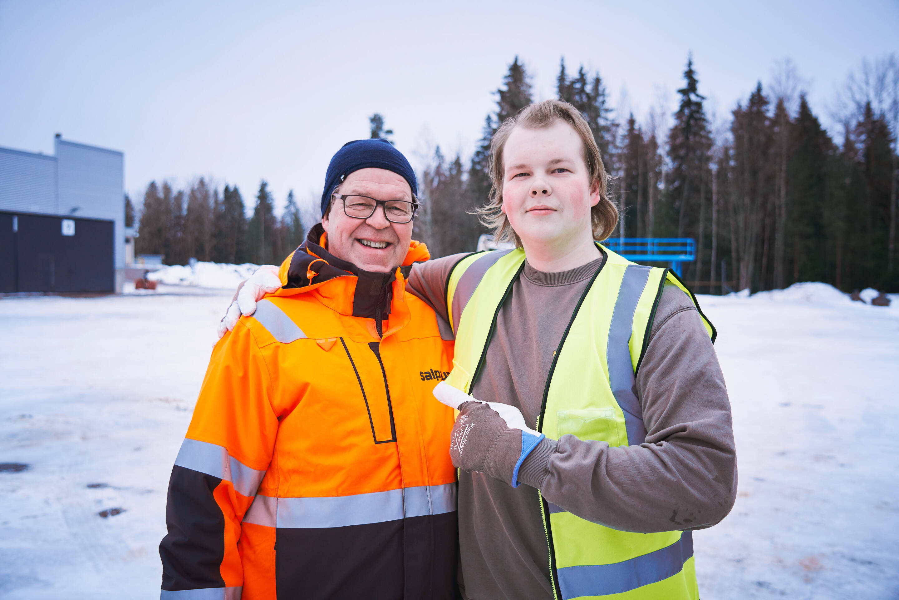 Kaksi ihmistä on ulkona kylmällä säällä. Toisella on yllään oranssi turvatakki ja silmälasit, hymyillen käsi toisella. Toisella henkilöllä on yllään keltainen turvaliivi, ja hänellä on paperilappu. Lumi peittää maan ja puut ovat taustalla.