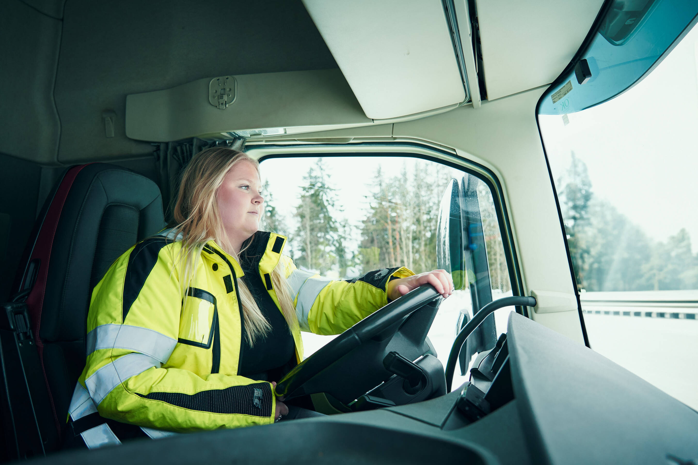 Pitkähiuksinen henkilö, jolla on yllään keltainen näkyvä takki, ajaa suurta kuorma-autoa. Kuorma-auton ohjaamon sisäpuoli näkyy, ja ulkotie on puiden reunustama.
