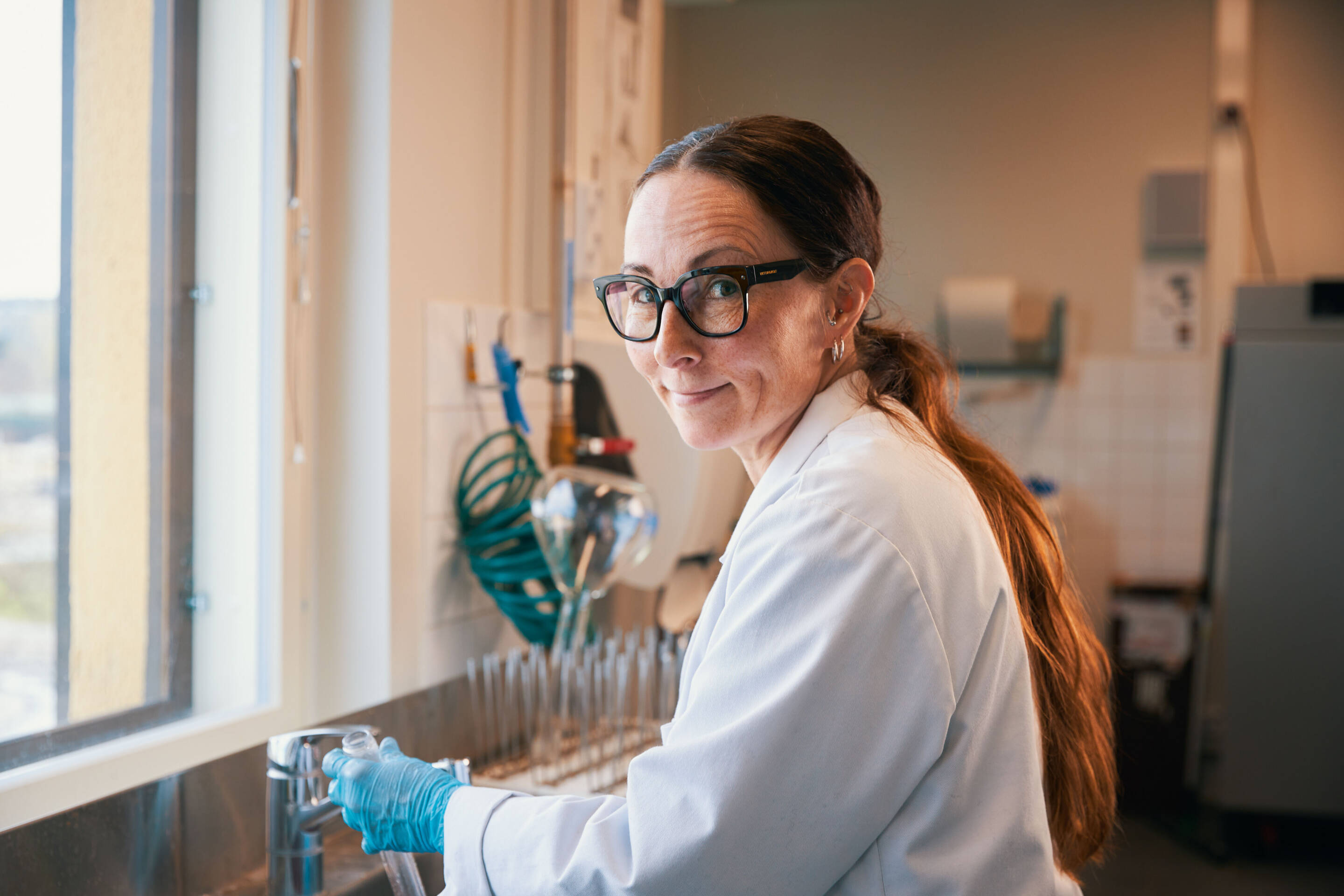 Pitkähiuksinen ja lasillinen henkilö hymyilee työskennellessään laboratoriossa. Heillä on yllään valkoinen laboratoriotakki ja siniset hanskat, ja he seisovat tiskin vieressä varusteineen. Taustalla näkyy ikkuna.