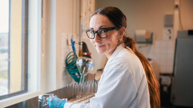 Pitkähiuksinen ja lasillinen henkilö hymyilee työskennellessään laboratoriossa. Heillä on yllään valkoinen laboratoriotakki ja siniset hanskat, ja he seisovat tiskin vieressä varusteineen. Taustalla näkyy ikkuna.