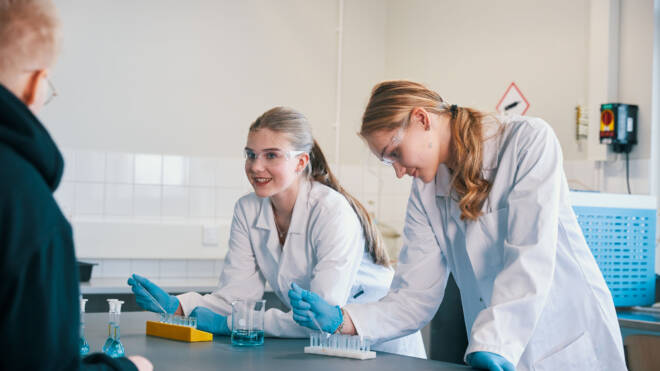 Kaksi ihmistä laboratoriotakissa ja suojalaseissa työskentelee laboratoriopenkillä. Toisessa on koeputkiteline ja toisessa muistikirja. He näyttävät keskustelevan jonkun kanssa kameran ulkopuolella. Laboratorion pöydällä on erilaisia tieteellisiä laitteita.