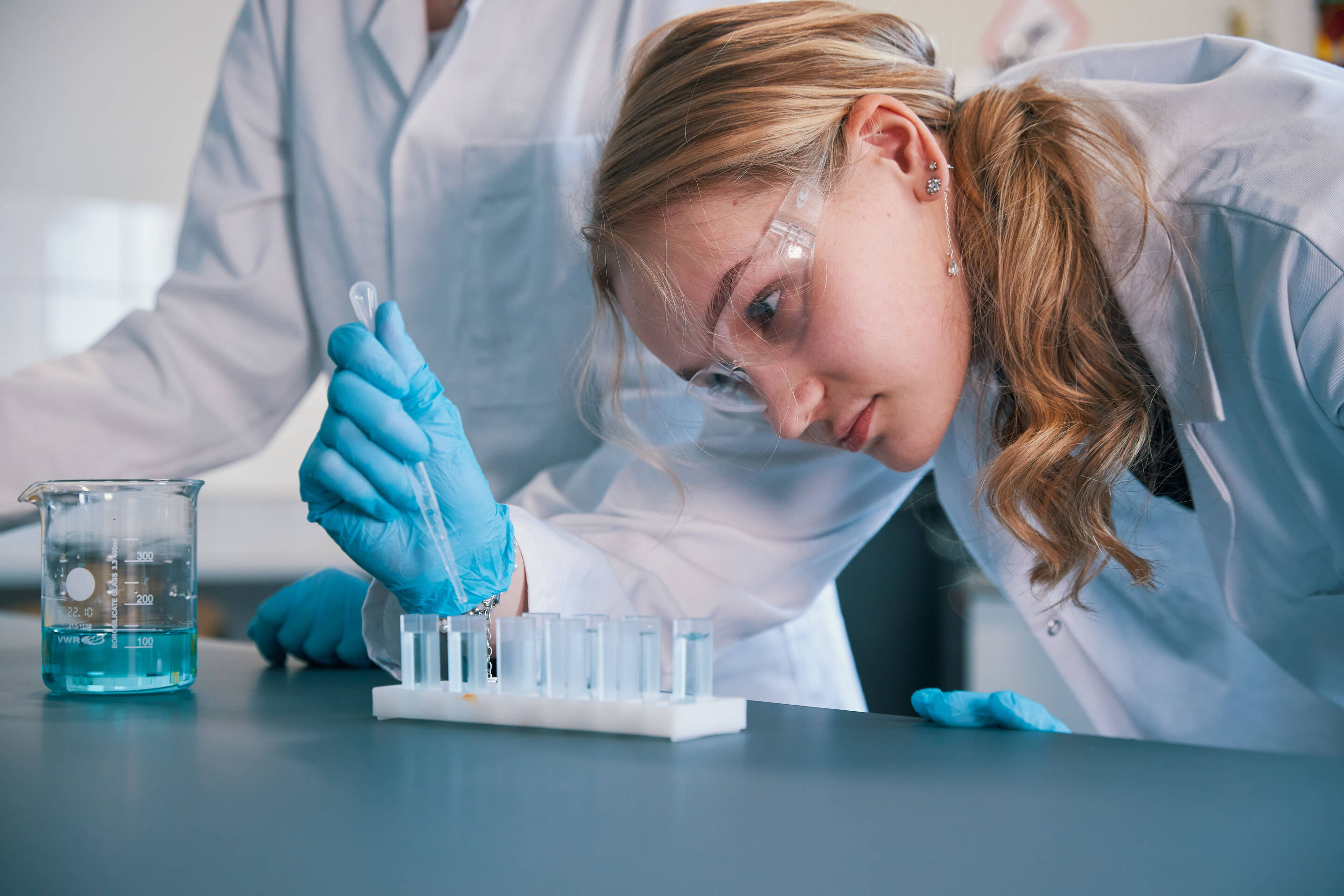 Nainen, jolla on suojalasit ja siniset hanskat, pipetoi nestettä varovasti koeputkeen laboratorioympäristössä. Toinen henkilö laboratoriotakissa seisoo lähellä. Pöydällä on sinistä nestettä sisältävä dekantterilasi. Taustalla näkyy tiedelaitteita.