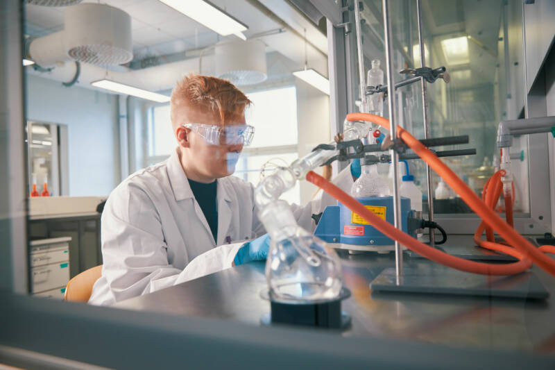 Suojalaseja ja laboratoriotakkia käyttävä henkilö suorittaa kokeen laboratoriossa. Niitä ympäröivät tieteelliset laitteet, kuten putket, lasitavarat ja pyörivä sekoitin. Laboratorio on hyvin valaistu moderneilla järjestelyillä.