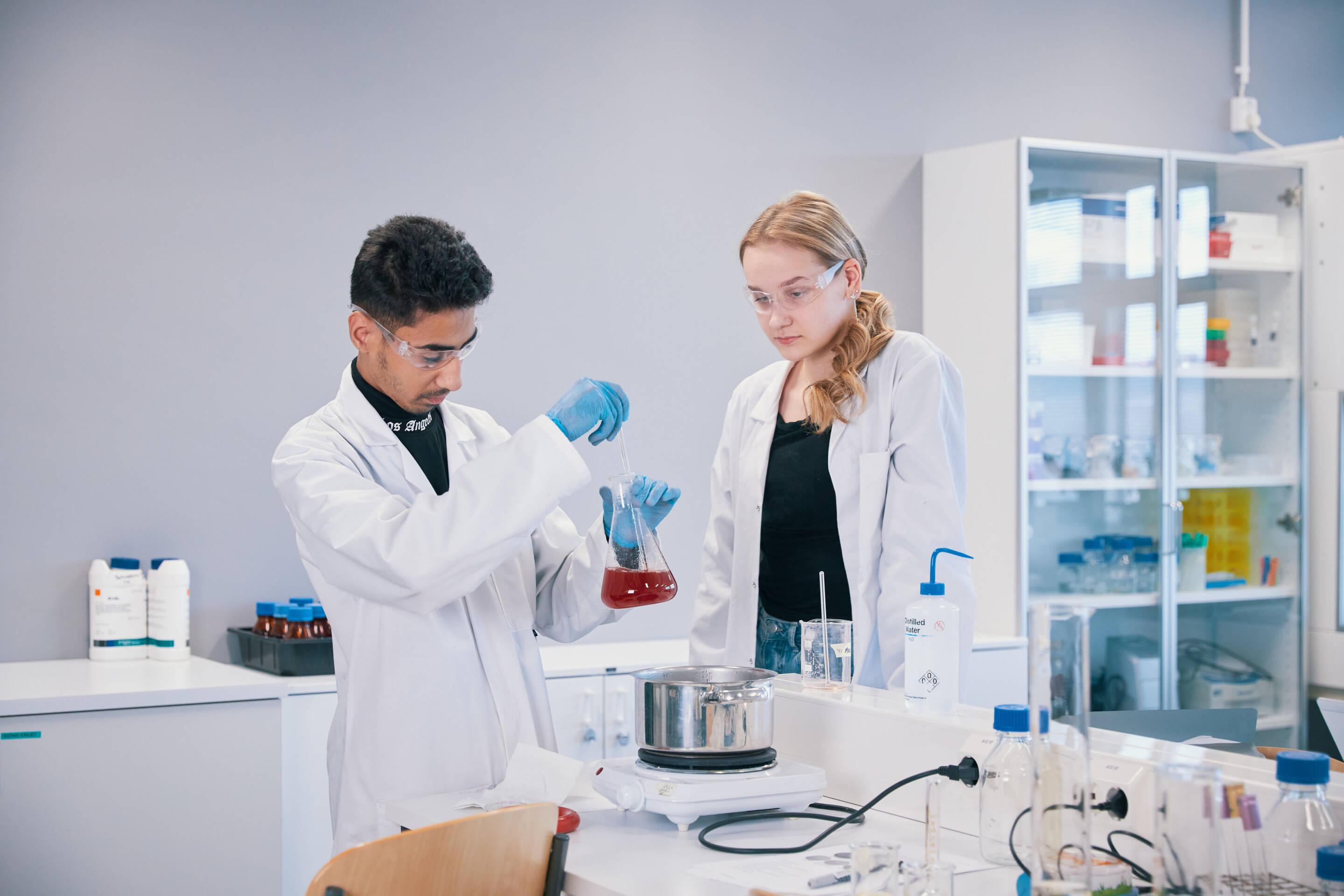 Kaksi tiedemiestä laboratoriossa tekevät kokeen. Toisella on pullo punaisella nesteellä käsineissä ja suojalaseissa. Toinen tarkkailee silmälaseja päällään. Taustalla on hyllyt laboratoriovälineillä.