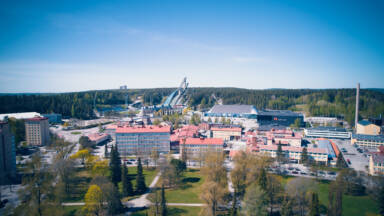 Ilmakuva kaupunkikuvasta, jonka taustalla on suuri mäkihyppyrakenne. Alla on punakattoisia rakennuksia vihreiden puiden ja ruohoalueiden ympäröimänä kirkkaan sinisen taivaan alla.