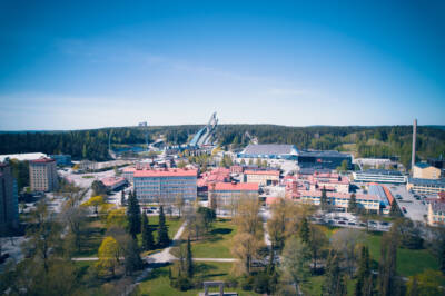 Ilmakuva kaupunkikuvasta, jonka taustalla on suuri mäkihyppyrakenne. Alla on punakattoisia rakennuksia vihreiden puiden ja ruohoalueiden ympäröimänä kirkkaan sinisen taivaan alla.