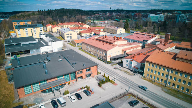 Ilmakuva pienestä kaupungista, jossa on punaisia ja beige-kattoisia rakennuksia. Autot pysäköidään suorakaiteen muotoisten rakenteiden reunustaman kadun varrelle. Puut ja mäkiset maisemat näkyvät taustalla osittain pilvisen taivaan alla.