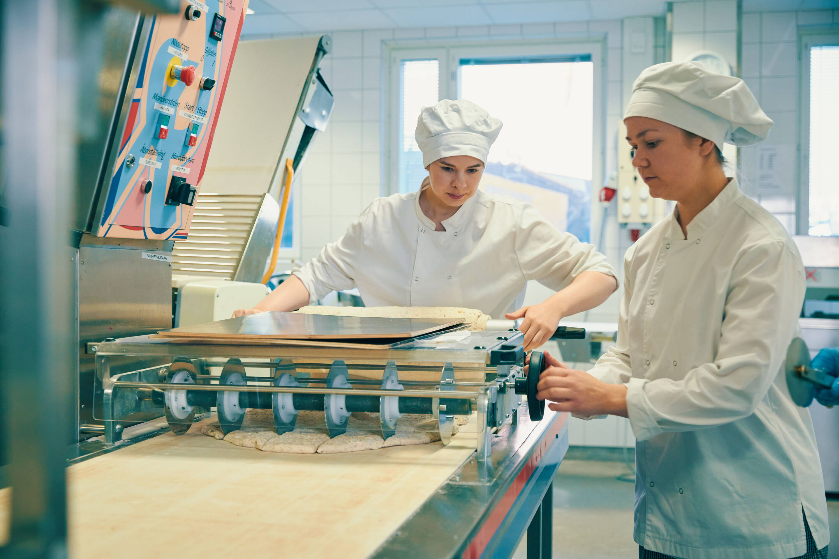 Kaksi leipuria valkoisissa univormuissa ja hatuissa työskentelevät yhdessä leipomokeittiössä. Toinen kaulii taikinaa koneella ja toinen säätelee sitä. Niitä ympäröivät leivontalaitteet ja taustalla oleva ikkuna päästää sisään luonnonvaloa.