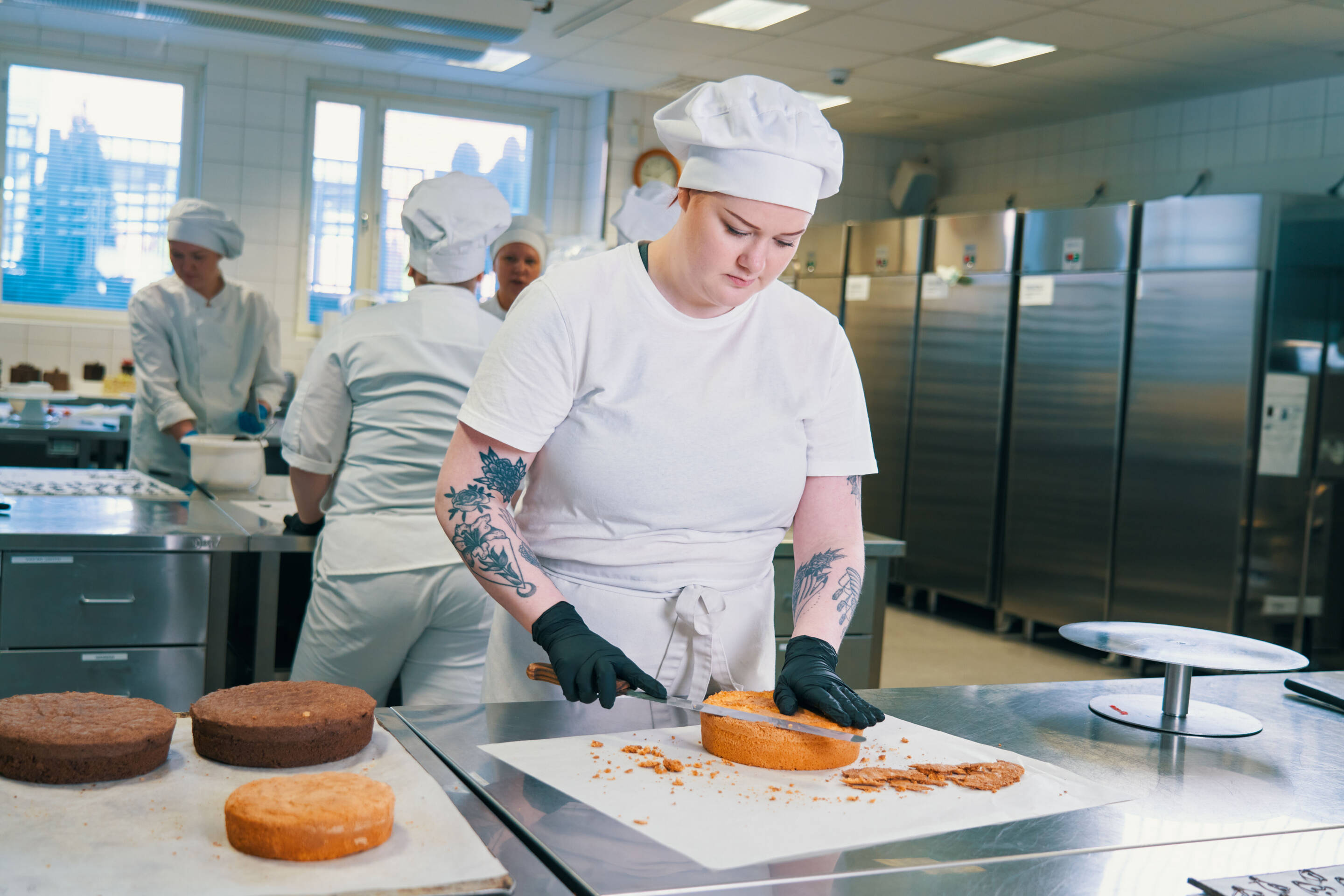 Keskittynyt kondiittori, jolla on valkoinen univormu ja lippalakki, koristaa kakun ammattikeittiössä. Työtila on siisti, taustalla näkyy muita kokkeja valmistamassa jälkiruokia. Puhtaat ruostumattomasta teräksestä valmistetut pinnat ja työkalut näkyvät.