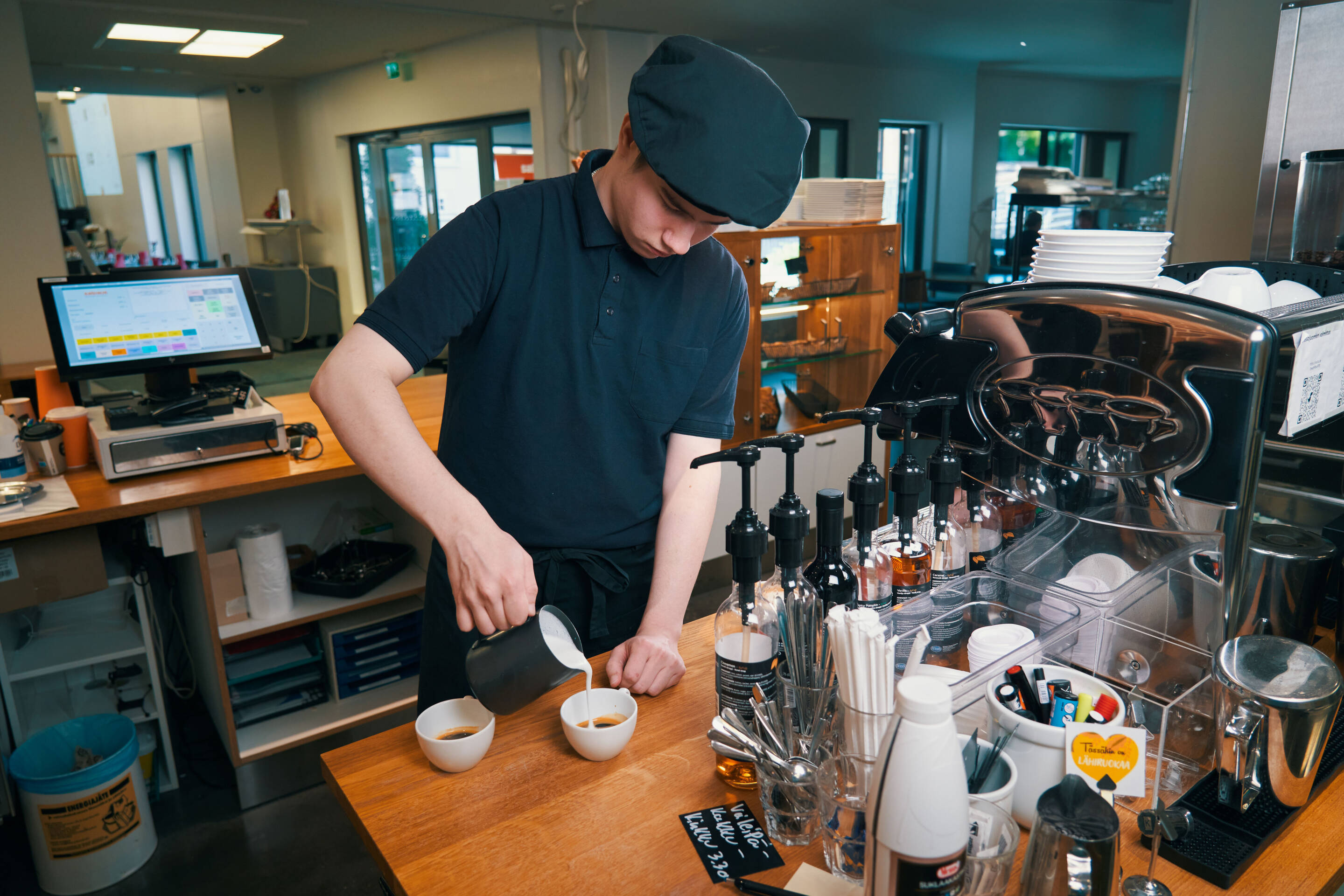 Mustassa työasussa oleva barista kaataa maitoa kahteen kahvikuppiin puisen tiskin takana. Lähistöllä on siirappipulloja, kahvinkeitin ja kassakone. Taustalla on hyllyjä ja ruostumattomasta teräksestä valmistettuja laitteita.
