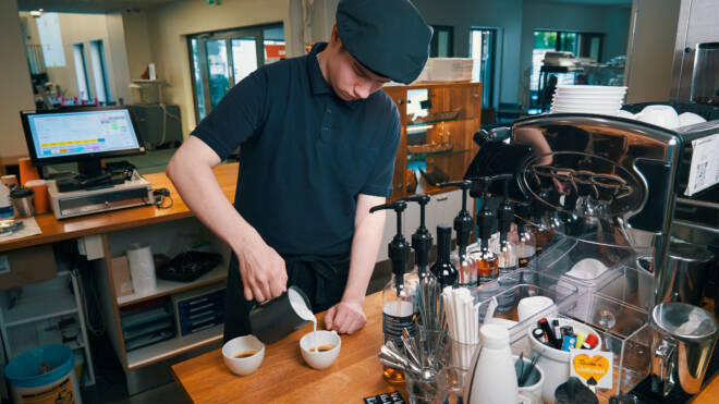 Mustassa työasussa oleva barista kaataa maitoa kahteen kahvikuppiin puisen tiskin takana. Lähistöllä on siirappipulloja, kahvinkeitin ja kassakone. Taustalla on hyllyjä ja ruostumattomasta teräksestä valmistettuja laitteita.