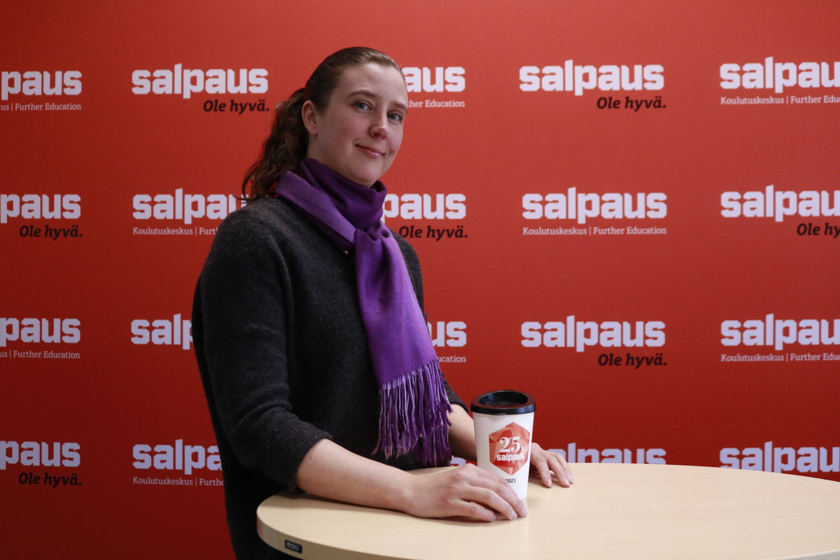Pitkät, taakse sidotut hiukset omaava henkilö, jolla on yllään tumma neulepusero ja violetti huivi, seisoo pöydän ääressä pitäen edessään kahvikuppia. Oranssissa taustassa on Salpauksen logoja ja teksti ole hyvä.