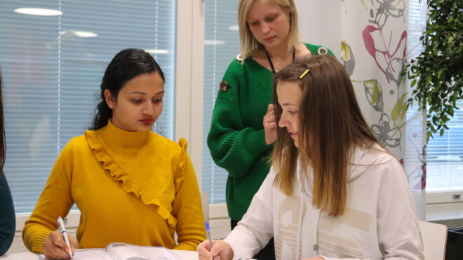 Kaksi henkilöä istuu pöydän ääressä muistivihkojen kanssa kirjoittamassa ja yksi seisomassa ja ohjaamassa.