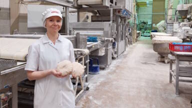 Valkoiseen leipuritakkiin ja päähineeseen pukeutunut henkilö seisoo leipomotehtaassa hymyillen ja pitelee kahta leipätaikinaa. Taustalla näkyvät teolliset leipomolaitteet ja jauhoinen lattia.
