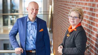 Kaksi hymyilevää aikuista, toinen siniseen pukuun pukeutunut mies ja toinen silmälaseihin ja oranssiin huiviin pukeutunut nainen, seisoo sisätiloissa tiiliseinän ja lasikaiteen vieressä kirkkaasti valaistussa modernissa rakennuksessa.