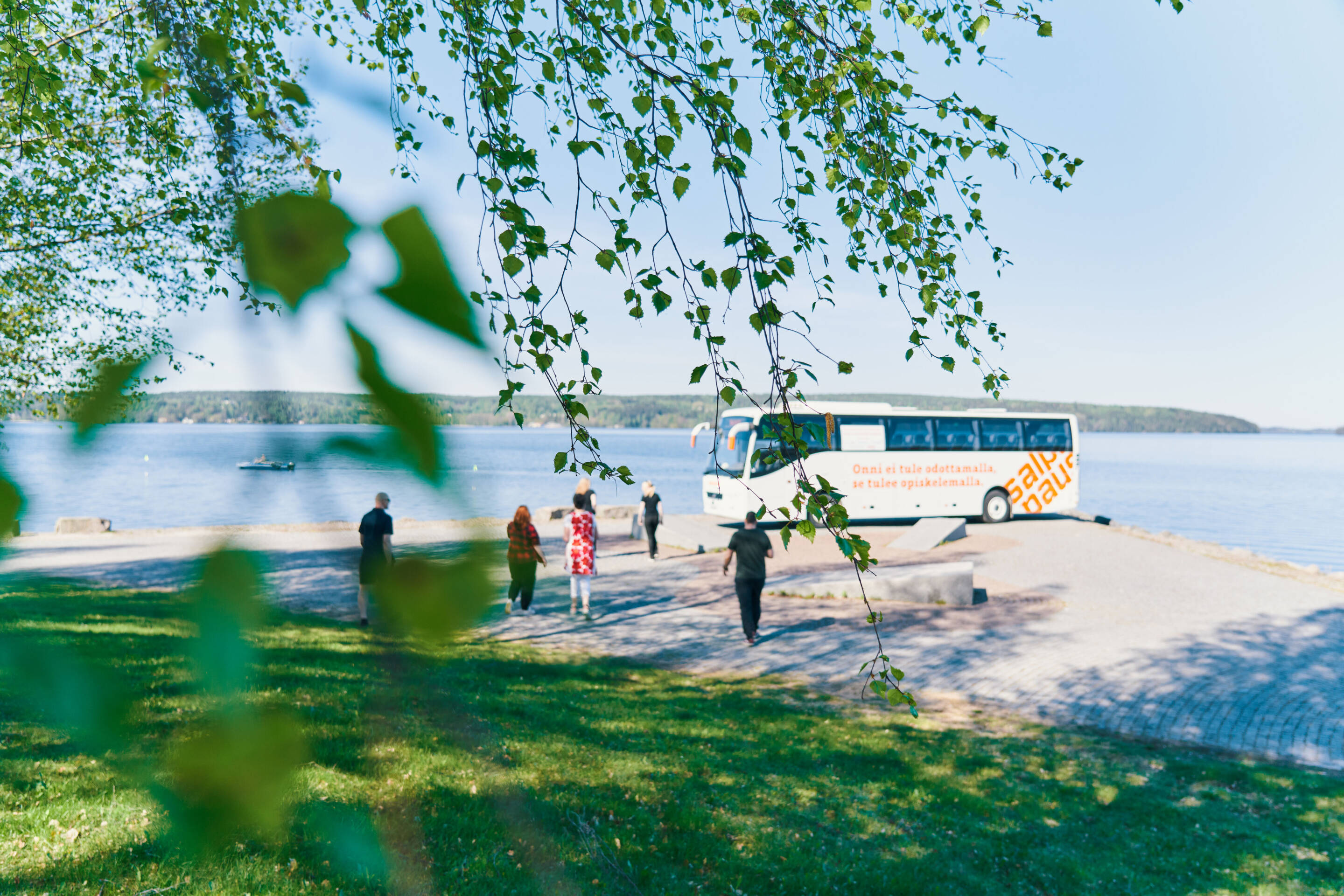 Bussi on pysäköity rauhallisen järvenrannan lähelle, jota kohti kävelee muutama ihminen. Läheisen puun vihreät lehdet kehystävät etualalla, ja taivas on kirkas ja sininen heijastaen järvien pintaa.