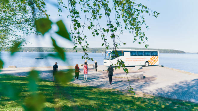 Bussi on pysäköity rauhallisen järvenrannan lähelle, jota kohti kävelee muutama ihminen. Läheisen puun vihreät lehdet kehystävät etualalla, ja taivas on kirkas ja sininen heijastaen järvien pintaa.