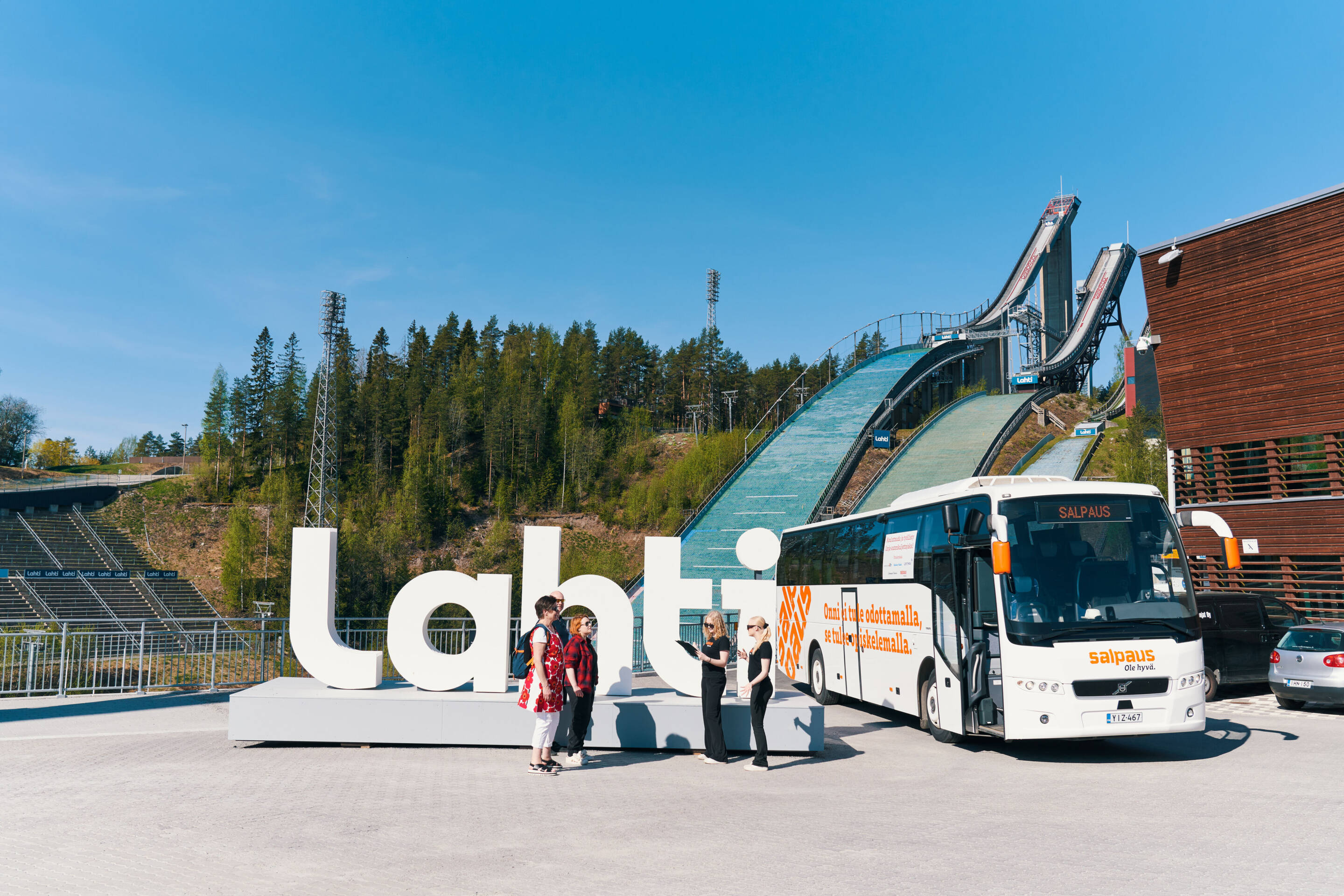 Turistit seisovat suuren Lahti-kyltin ääressä hyppyrimäkien edessä Lahdessa. Valkoinen bussi on pysäköity lähelle kirkkaan sinisen taivaan alla..