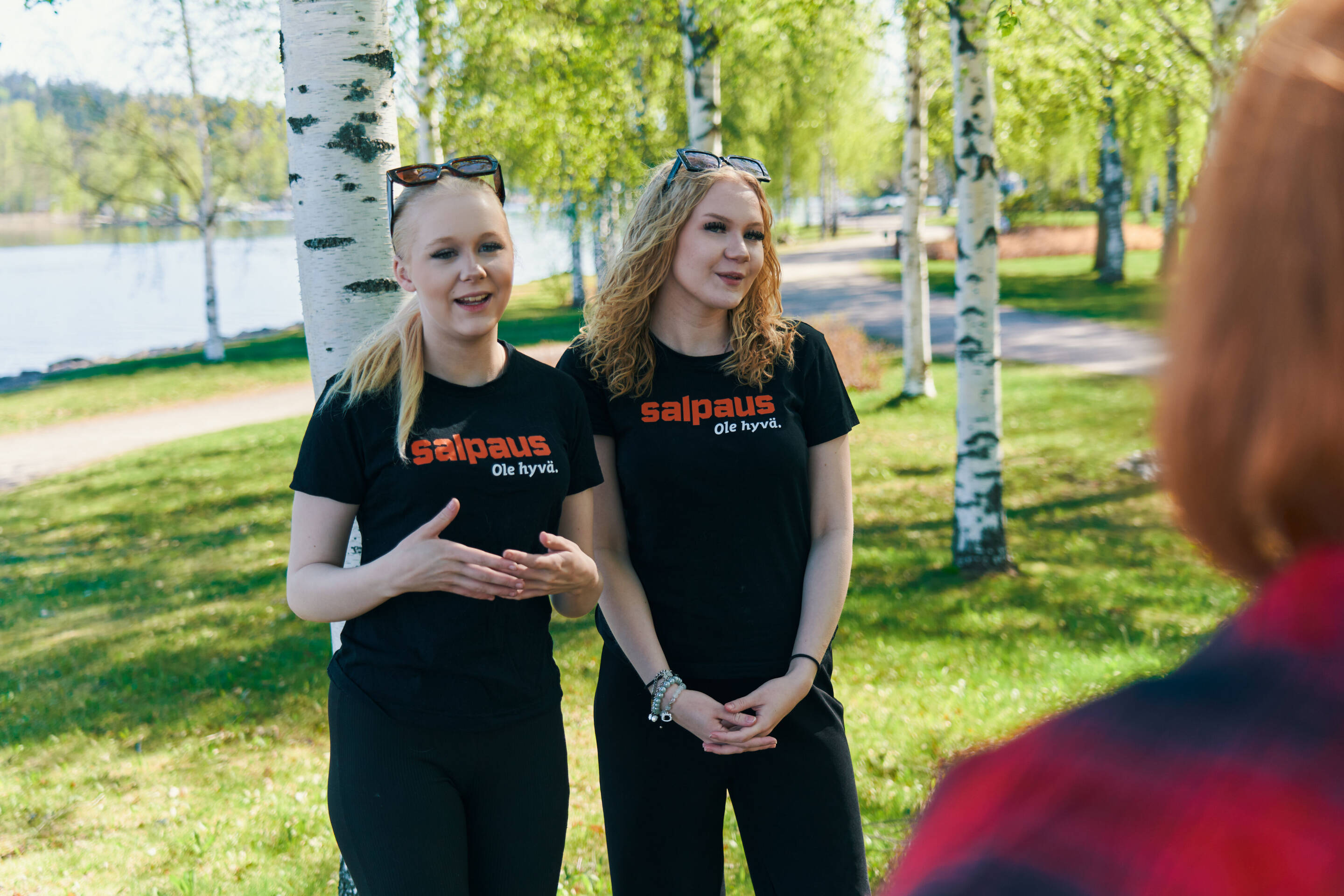 Kaksi mustassa Salpaus-paidassa pukeutunutta ihmistä seisoo puistossa koivujen ja vesistön taustalla. He puhuvat jollekulle kameran ulkopuolella auringon paistaessa kirkkaasti.