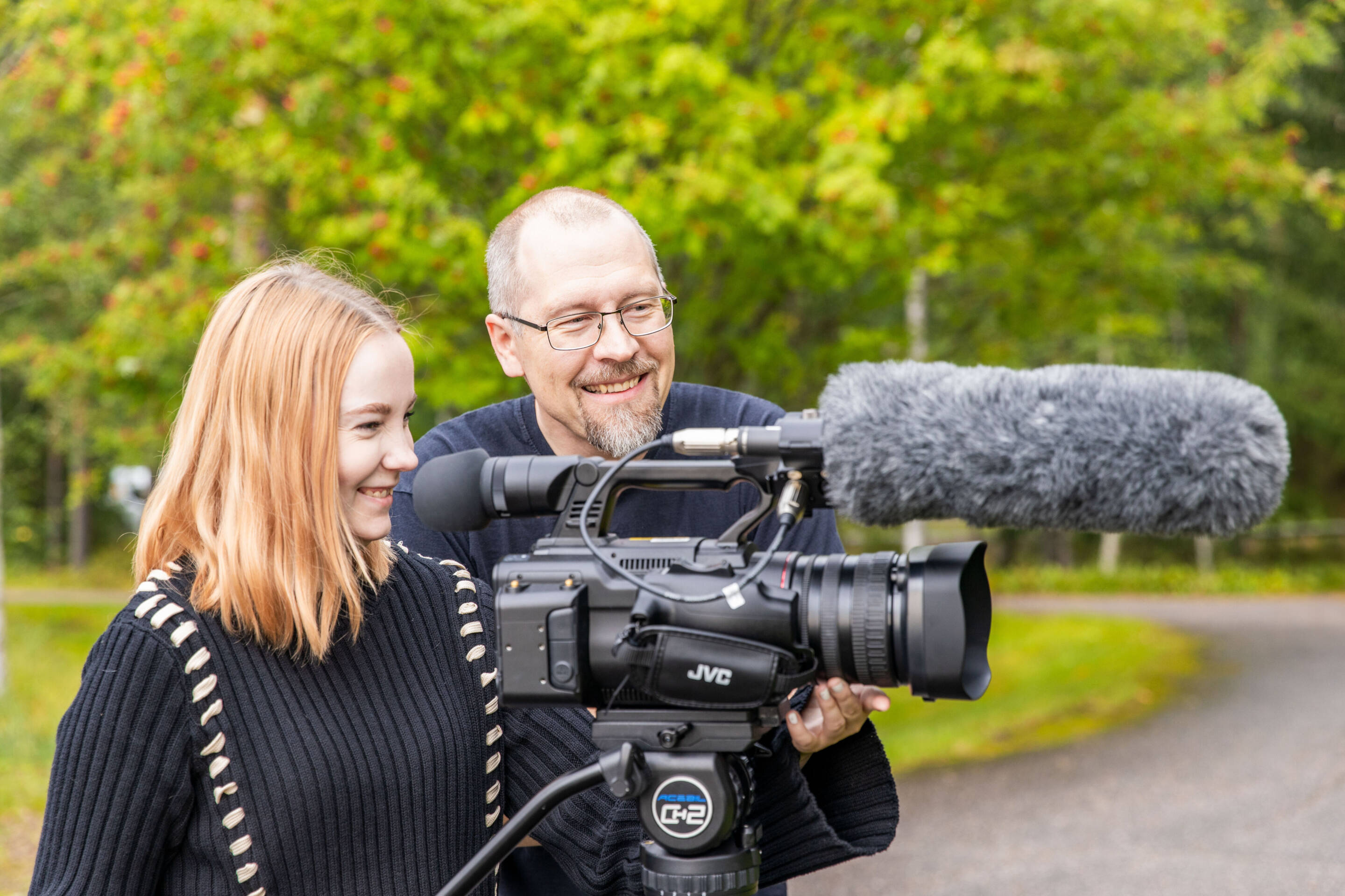 Nuori nainen ja mies ovat ulkona hymyillen ja seisovat ammattivideokameran takana jalustalla. Kamerassa on iso mikrofoni. He näyttävät kuvaavan luonnollisessa ympäristössä vihreiden puita taustalla.