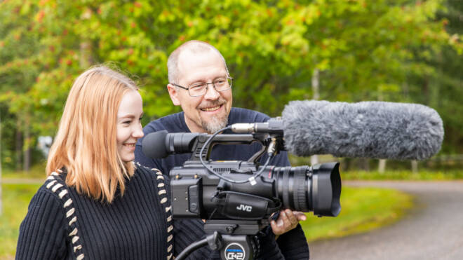 Nuori nainen ja mies ovat ulkona hymyillen ja seisovat ammattivideokameran takana jalustalla. Kamerassa on iso mikrofoni. He näyttävät kuvaavan luonnollisessa ympäristössä vihreiden puita taustalla.