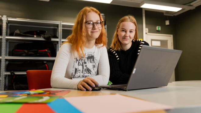 Kaksi ihmistä istuu pöydän ääressä kannettavalla tietokoneella. Yksi silmälaseissa käyttää hiirtä, kun taas toinen hymyilee kameralle. Taustalla värikäs paperi ja siisti huone, jossa on hylly.