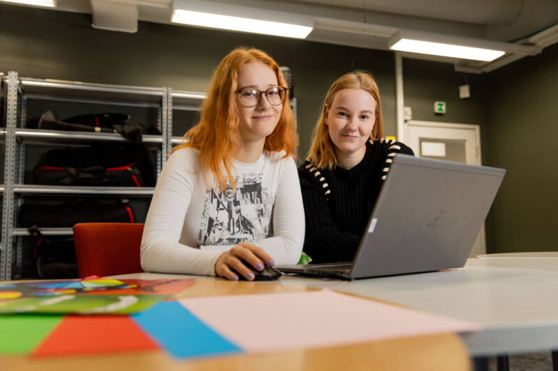 Kaksi ihmistä istuu pöydän ääressä kannettavalla tietokoneella. Yksi silmälaseissa käyttää hiirtä, kun taas toinen hymyilee kameralle. Taustalla värikäs paperi ja siisti huone, jossa on hylly.