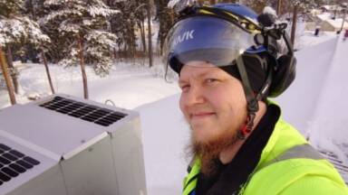 Henkilö neonkeltaisessa takissa ja suojakypärässä kuulosuojaimilla hymyilee kameralle seisoessaan lumisella katolla LVI-yksikön vieressä. Taustalla näkyy mäntyjä ja lumen peittämää maisemaa.