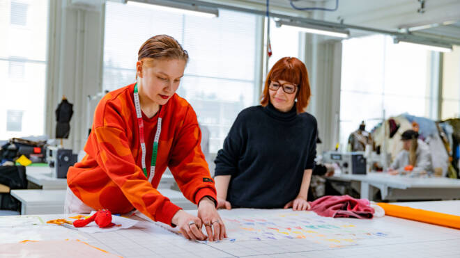 Kaksi naista työskentelee suunnittelustudiossa. Toinen punaisessa puserossa keskittyy kangastilkkujen asettamiseen pöydälle, kun taas toinen mustassa kelloa lähellä. Ompelukoneet ja materiaalit näkyvät taustalla.