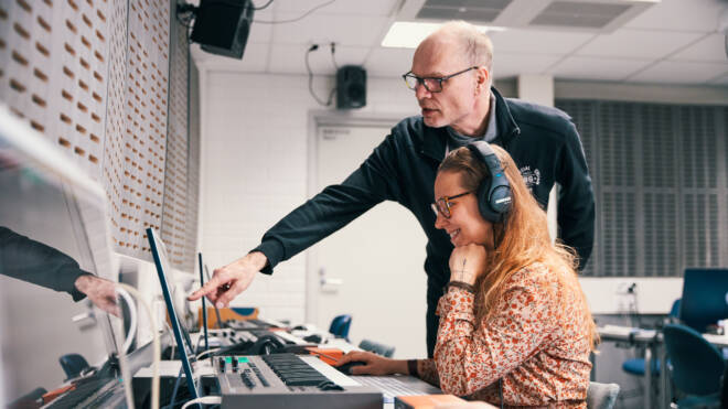 Mies osoittaa tietokoneen näyttöä ja nainen, jolla on kuulokkeet, istuu sen edessä hymyillen. He ovat huoneessa, jossa on äänilaitteet ja näppäimistöt.
