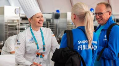 Hymyilevä leipuri-kondiittori valkoisessa työasussa ja korkeassa hatussa keskustelee kahden sinisessä "Support Team Finland" -takissa pukeutuneen ihmisen kanssa. Taustalla on ammattikeittiö.