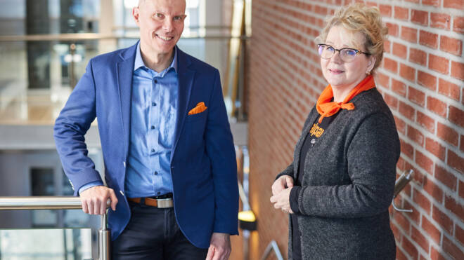 Mies sinisessä puvussa ja oranssissa taskuruudussa seisoo naisen vieressä harmaassa neuletakissa, jossa on oranssi huivi. He ovat sisäportaikoissa tiiliseinätaustalla hymyillen kameralle.