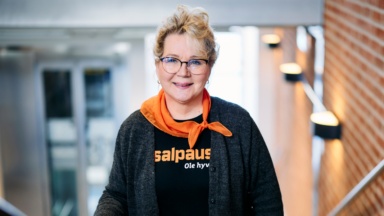 Nainen, jolla on vaaleat hiukset ja silmälasit, yllään oranssi huivi, musta neuletakki ja musta tekstillinen paita, seisoo portaikossa, jossa on tiiliseinät ja modernit valot.