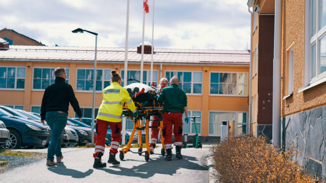 Päivystyshenkilöstö pyöräilee potilasta paareilla kohti rakennusta. Joukkue on pukeutunut kirkkaisiin univormuihin, ja kohtaus sijoittuu pysäköintialueelle, jossa on autoja ja iso rakennus taustalla.