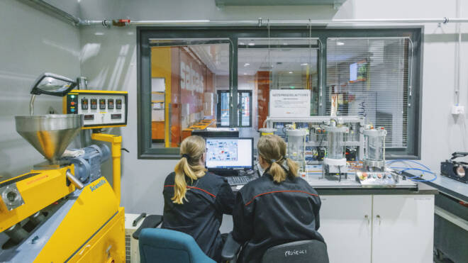 Kaksi mustaan takkiin pukeutunutta ihmistä istuu tietokoneen ääressä modernissa laboratoriossa. He katsovat poispäin ja valvovat laitteita, jotka on kytketty keltaiseen koneeseen. Lasi-ikkuna näyttää toisen huoneen.