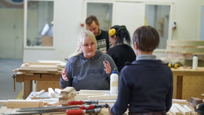 Nainen eleilee puhuessaan toiselle henkilölle puuntyöstöliikkeessä. Pöydällä näkyy erilaisia työkaluja ja puuosia. Taustalla työskentelee kaksi muuta henkilöä. Ympäristö on kiireinen ja täynnä puuntyöstölaitteita.
