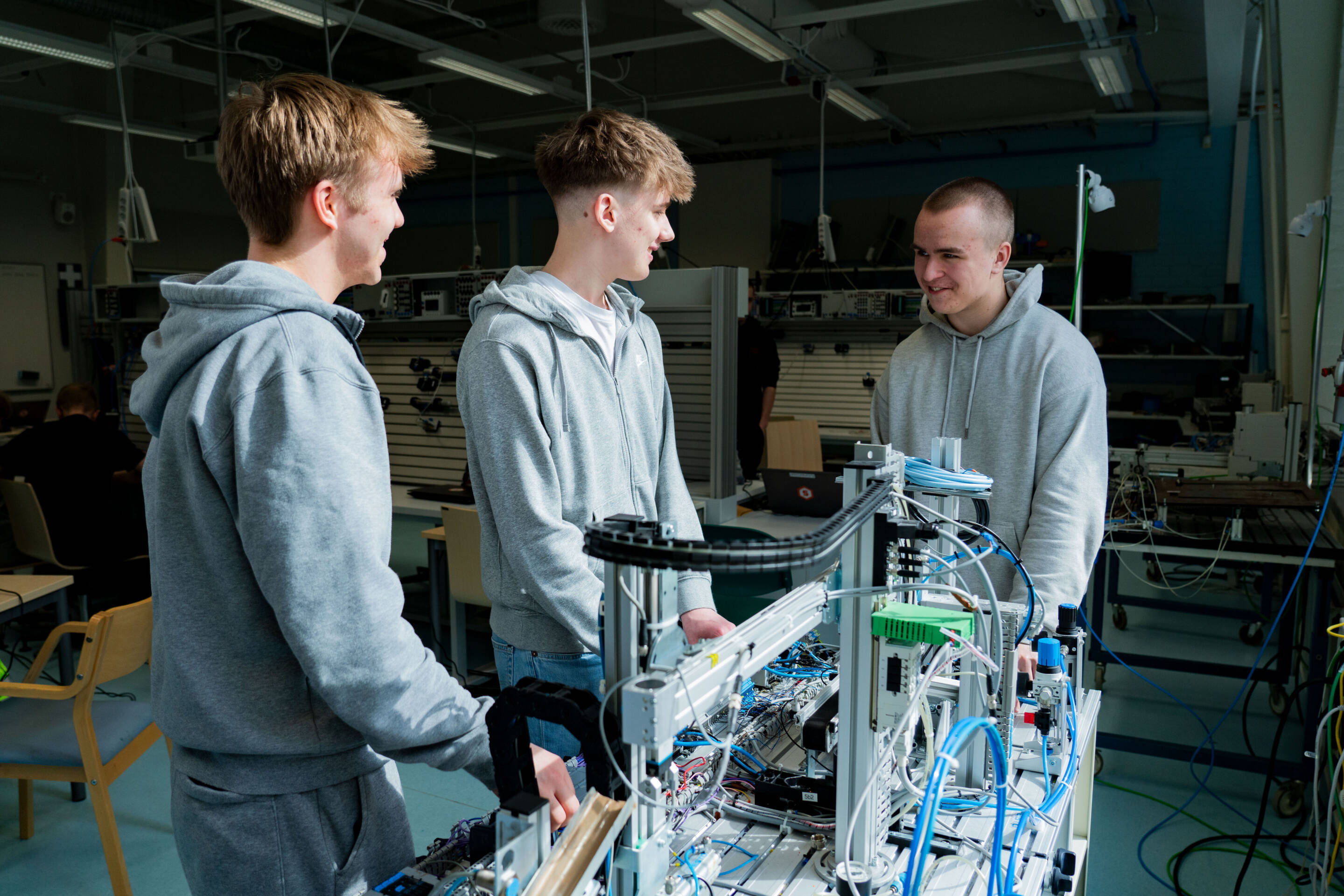 Kolme nuorta miestä työpajassa seisoo robotiikkaprojektin ympärillä hymyillen ja keskustelemassa. He käyttävät rentoja harmaita huppareita huoneessa, joka on täynnä elektronisia laitteita ja työkaluja.