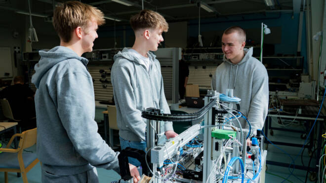 Kolme nuorta miestä työpajassa seisoo robotiikkaprojektin ympärillä hymyillen ja keskustelemassa. He käyttävät rentoja harmaita huppareita huoneessa, joka on täynnä elektronisia laitteita ja työkaluja.
