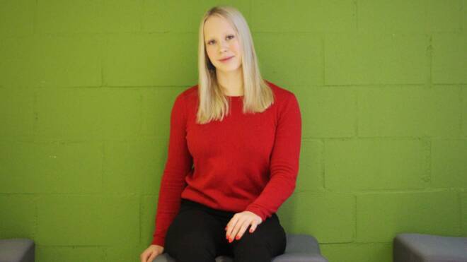 Nuori nainen, jolla on pitkät vaaleat hiukset, punainen pusero ja mustat housut yllään, istuu harmaalla kuutiolla vihreää tiiliseinää vasten.
