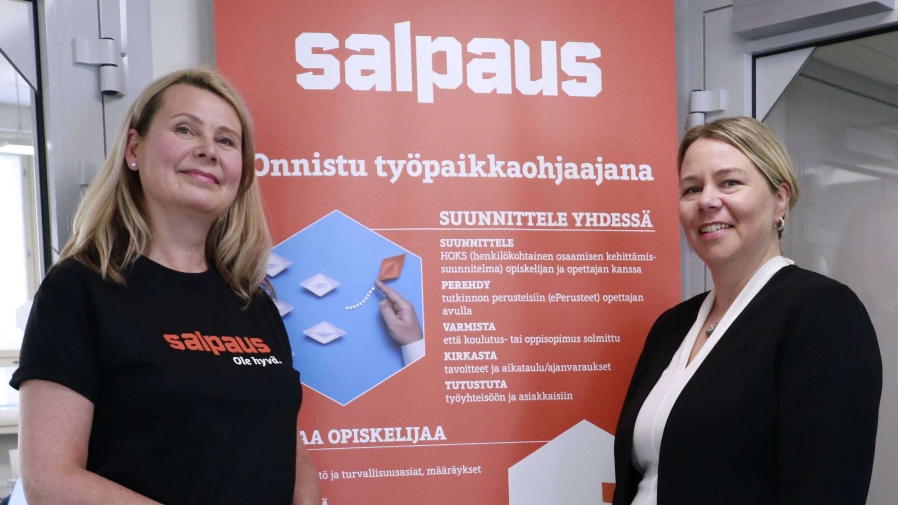 Kaksi tummiin vaatteisiin pukeutunutta naista seisovat ja hymyilevät oranssin roll upin edessä, jossa lukee Onnistu työpaikkaohjaajana.