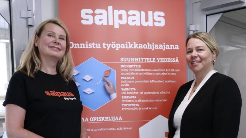 Kaksi tummiin vaatteisiin pukeutunutta naista seisovat ja hymyilevät oranssin roll upin edessä, jossa lukee Onnistu työpaikkaohjaajana.