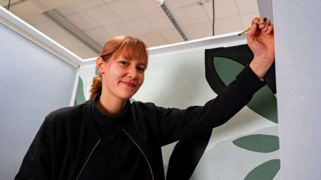 Ponihäntäinen henkilö hymyilee maalatessaan suuria lehtiä seinälle. Hän käyttää mustaa takkia ja pitää sivellintä oikeassa kädessään ja ojentaa kätensä seinää kohti. Taustalla näkyy katto valoineen.