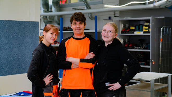 Kolme työasuihin pukeutunutta ihmistä seisoo yhdessä hymyillen. He käyttävät oransseja ja mustia asuja. Teollisuuden hyllyt ja laitteet näkyvät taustalla.