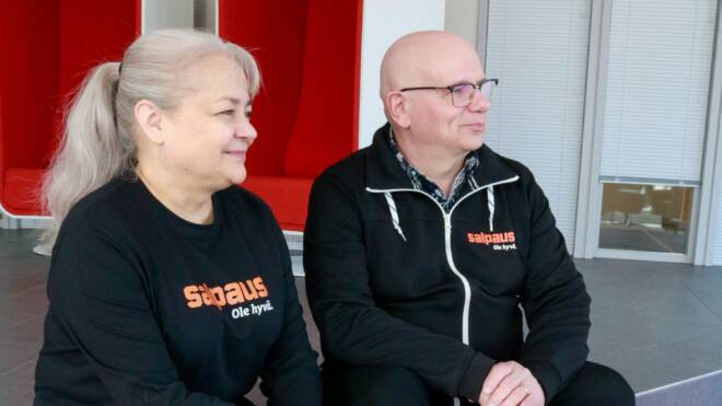 Kaksi ihmistä istuu vierekkäin sisätiloissa mustissa vaatteissa, joissa on Salpauksen logo. He ovat kasvot hieman oikealle hymyillen kasvoillaan.