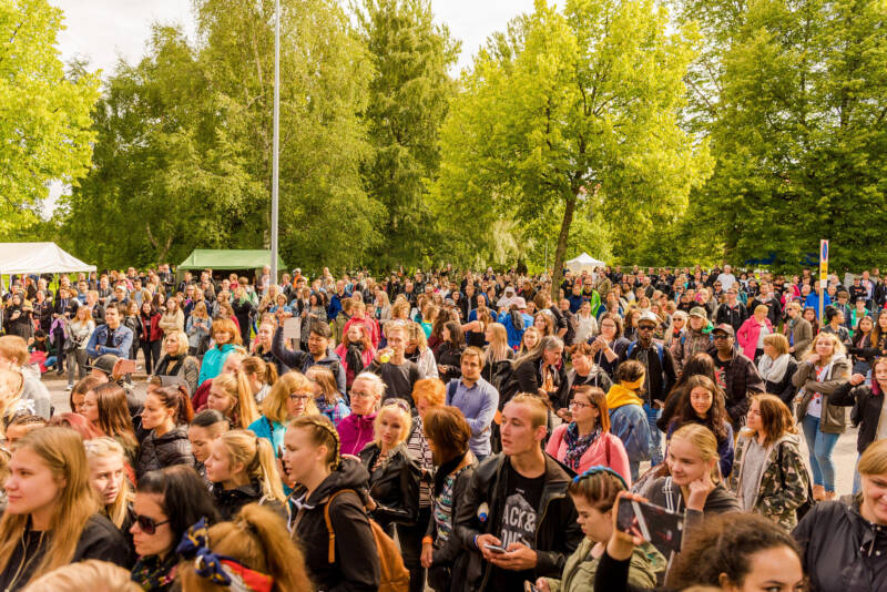Iso joukko ihmisiä ulkotapahtumassa. Taustalla näkyy muutamia telttakatoksia. Auringon säteet näkyvät taustalla olevissa puissa.